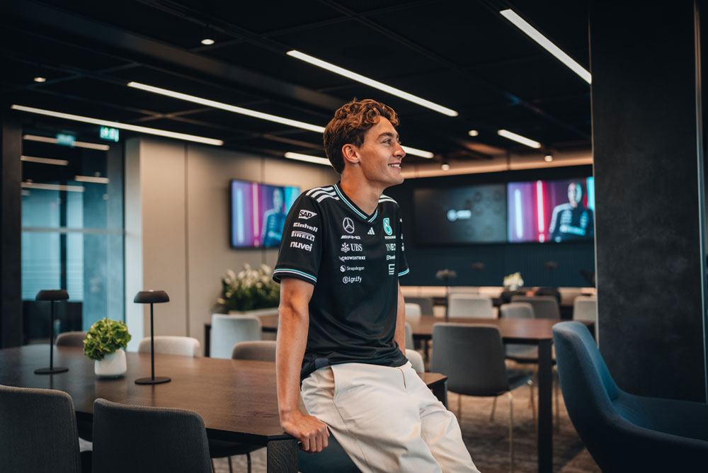 George Russell, coureur voor het Mercedes-AMG PETRONAS Formula One Team, zit ontspannen op een tafel in een moderne, stijlvolle teamruimte of kantooromgeving. Hij glimlacht en draagt het officiële teamshirt met zichtbare sponsorloco’s zoals Einhell, IWC en CrowdStrike. Op de schermen op de achtergrond is zijn afbeelding te zien.