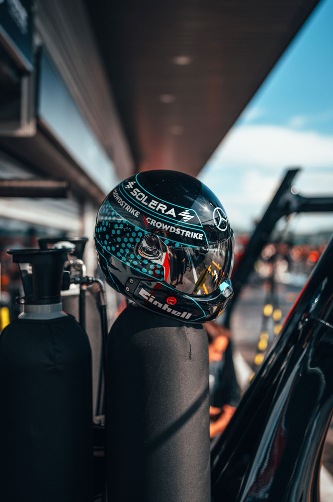 Een helm van het Mercedes-AMG PETRONAS Formula One Team met duidelijk zichtbare sponsorloco’s, waaronder Einhell, CrowdStrike en Solera, staat op een gasfles in de pitstraat. De reflectie in het vizier toont de omgeving van de garage.