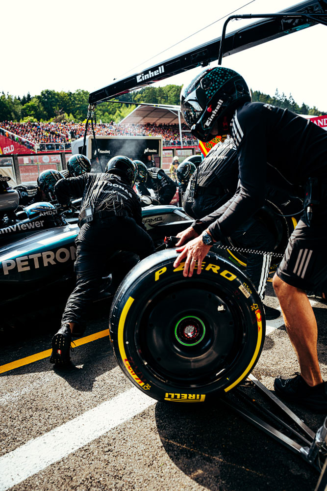 Monteurs van het Mercedes-AMG PETRONAS Formula One Team voeren een pitstop uit. Een teamlid bereidt een Pirelli-band voor terwijl anderen aan de auto werken. De Einhell-branding is zichtbaar op de apparatuur boven de auto.