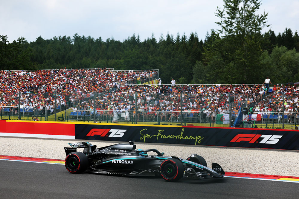 Een Mercedes-AMG PETRONAS Formula One-auto rijdt op het circuit van Spa-Francorchamps. Op de achtergrond zijn volle tribunes te zien met fans die internationale vlaggen zwaaien. Het Spa-Francorchamps-logo is zichtbaar op de barrière.