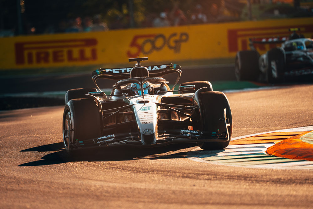 Een Mercedes-AMG PETRONAS Formula One-auto rijdt op een zonovergoten circuit tijdens een Grand Prix. De auto neemt met hoge snelheid een bocht terwijl een andere auto op de achtergrond te zien is.