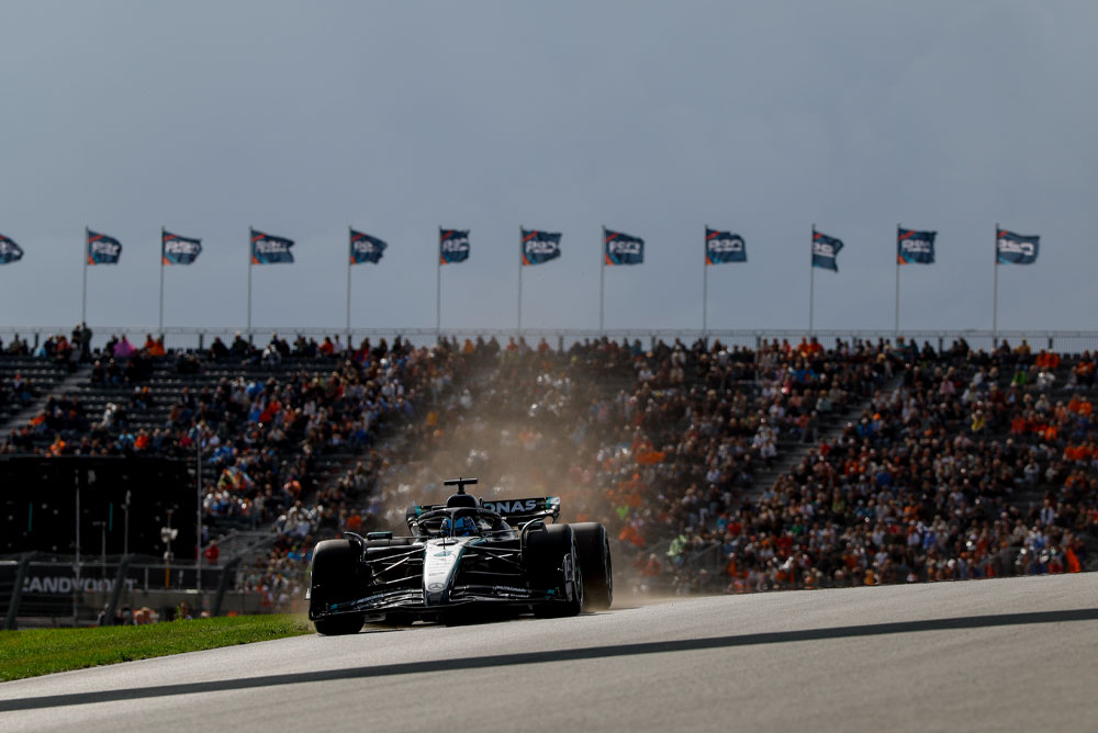 Een Mercedes-AMG PETRONAS Formula One-auto rijdt langs volle tribunes tijdens een race. Fans zwaaien met vlaggen op de achtergrond en creëren een levendige sfeer.